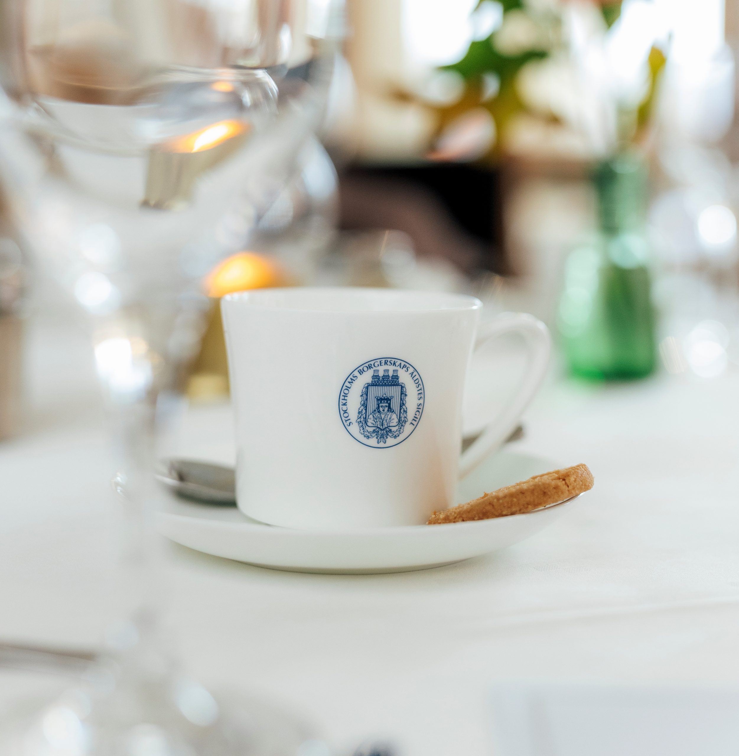 Närbild på en kopp med Borgerskapets logotyp och en kaka, omgiven av elegant dukade glas och blommor.