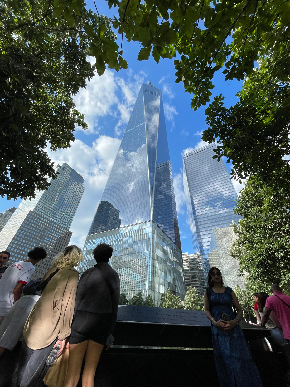 Studenter i New York framför One World Trade Center med skyskrapor och träd i bakgrunden.