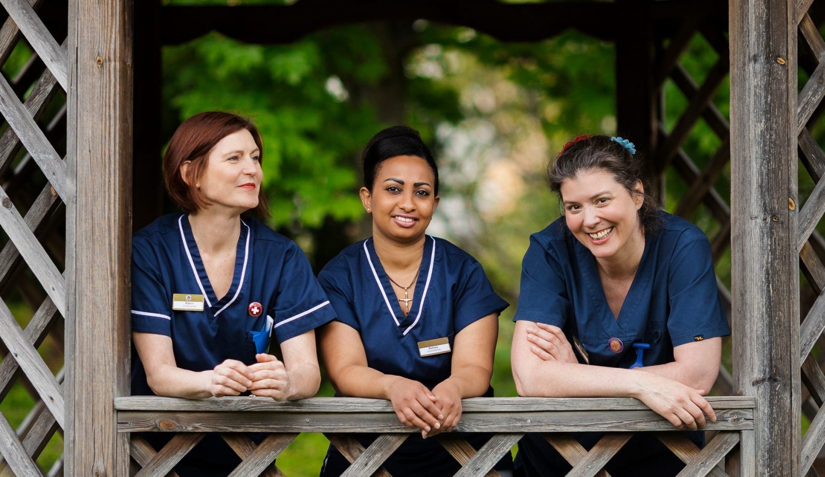 Tre vårdanställda i blå uniformer ler och samtalar utomhus i en träpaviljong omgiven av grönska.