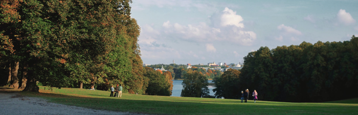 Vy över grönskande park med grön gräsmatta och personer som promenerar i parken med blå himmel ovanför.